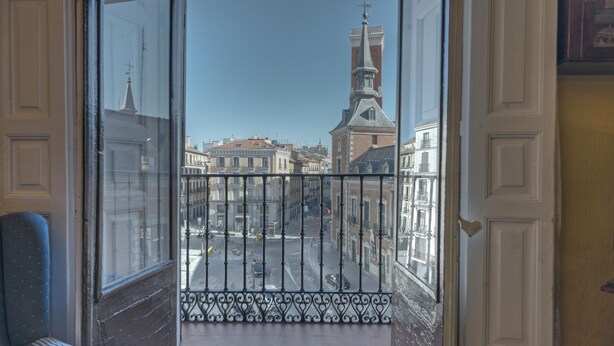Un balcone con ringhiera in ferro battuto che si affaccia su una strada con edifici e il campanile di una chiesa.