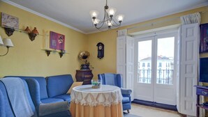 Un soggiorno con un divano blu, un tavolino con tovaglia, un lampadario, una sveglia e una vista all'esterno tramite una porta.