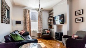 Een woonkamer met een zwarte leren bank, een grijze fauteuil, een ronde salontafel en een aan de muur bevestigde televisie.
