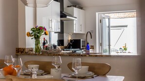Een moderne keuken met witte kastjes, een eethoek met een tafel gedekt voor twee personen, en uitzicht op de buitenlucht door een open deur.