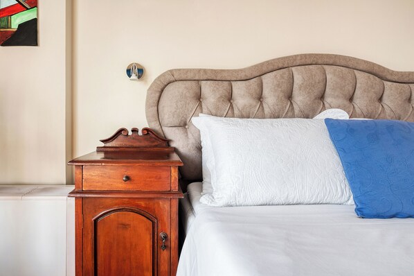 A bedroom with a tufted headboard, a wooden nightstand, a white bed with a blue throw blanket, and a wall-mounted light fixture.