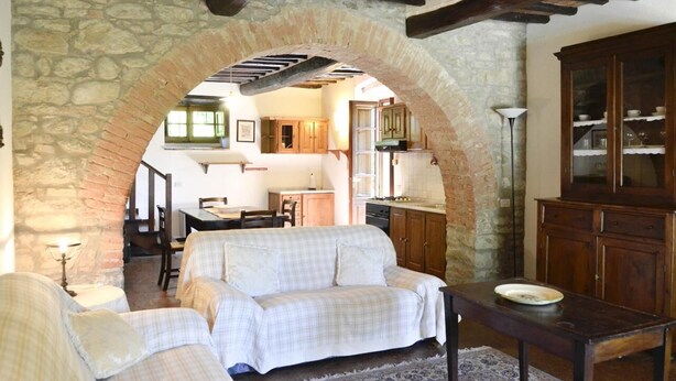 A cozy living room with a stone archway, a sofa, a wooden table, and a kitchen in the background.
