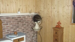 A room with a brick fireplace, a white vase on a metal stand, a small wooden cabinet, and a window with a view of the outside.
