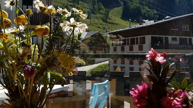Una ventana con vistas a un paisaje montañoso, una mesa de madera con un jarrón de flores y una silla.