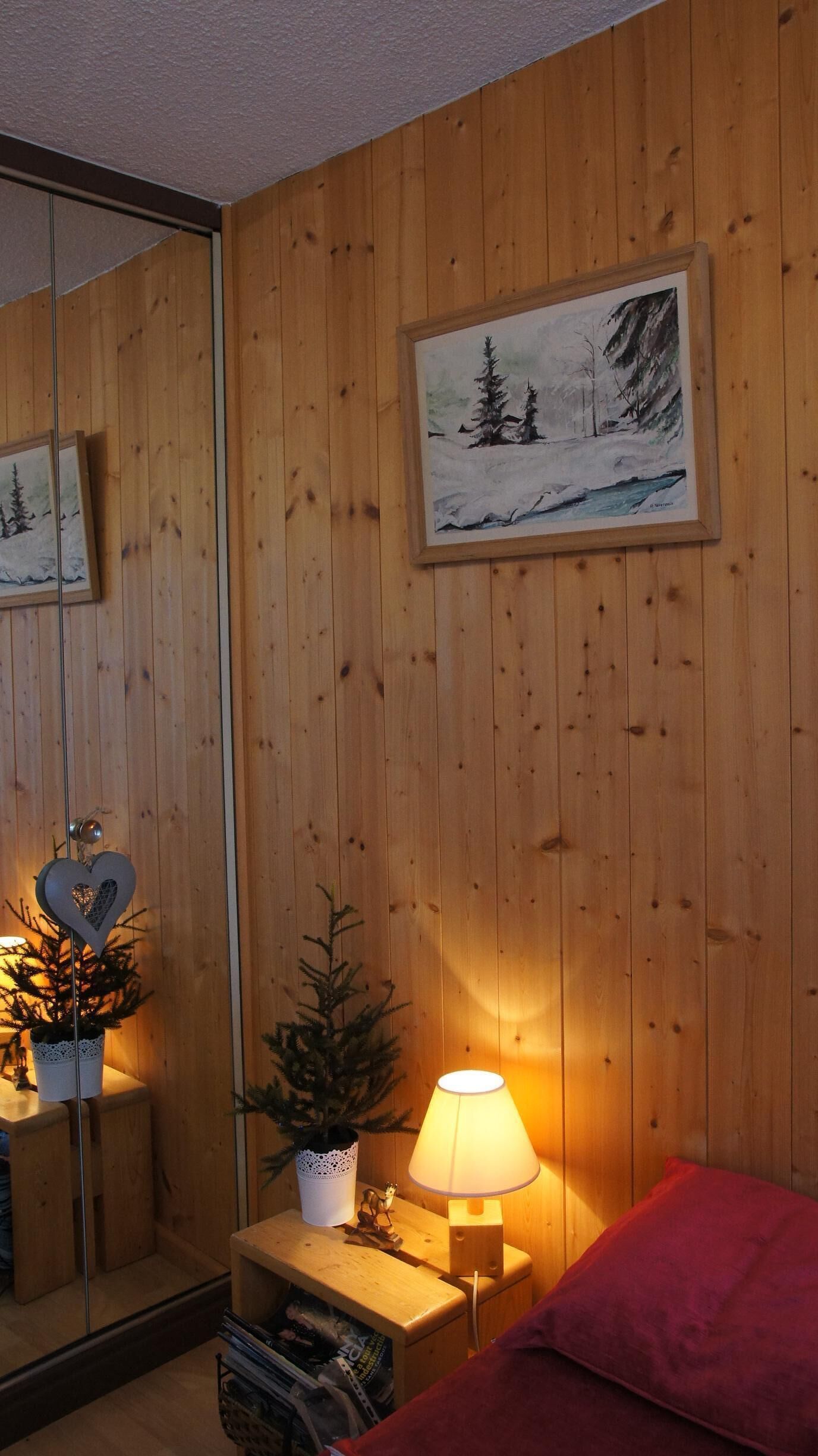 Quarto com paredes de madeira, um espelho, pintura de uma paisagem nevada, decoração em formato de coração, uma pequena mesa com uma planta e um abajur.