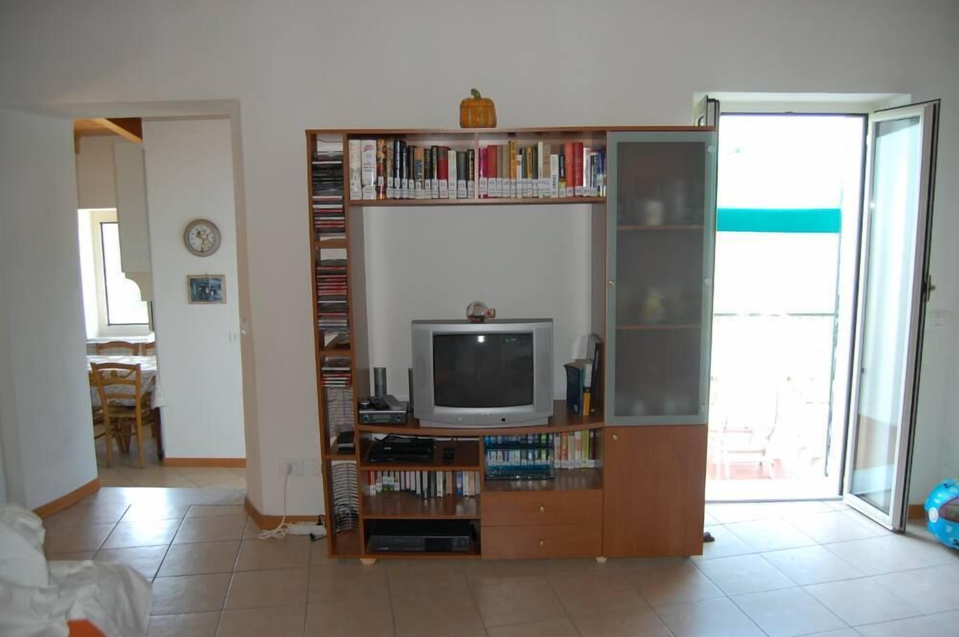 A living room with a television on a wooden stand, a bookshelf, and a dining area visible in the background.