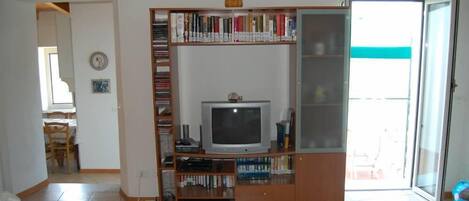 A living room with a television on a wooden stand, a bookshelf, and a dining area visible in the background.