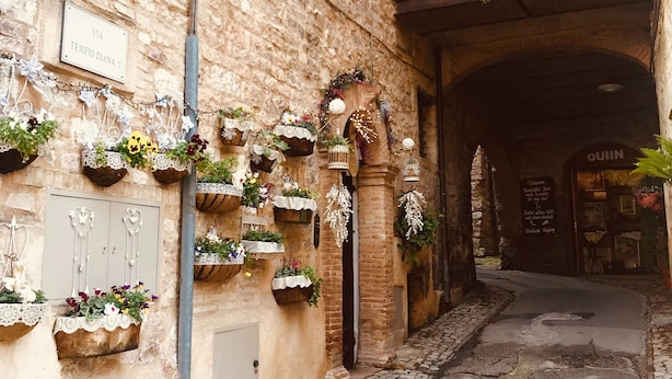 A narrow alley with a stone building, decorative hanging plants, and a sign reading "VIA TEMPIO DIANA".