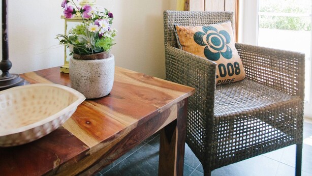 Een kamer met een houten tafel, een potplant, een rieten stoel met kussen en een raam met gordijnen.