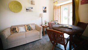 Een woonkamer met een beige bank, een houten eettafel en stoelen. Er staat een lamp, een wandmand en er hangen ingelijste foto's aan de muur.