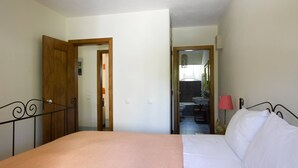 Een slaapkamer met een bed, een houten deur, een zichtbare badkamer door een open deur en een klein tafeltje met een lamp.
