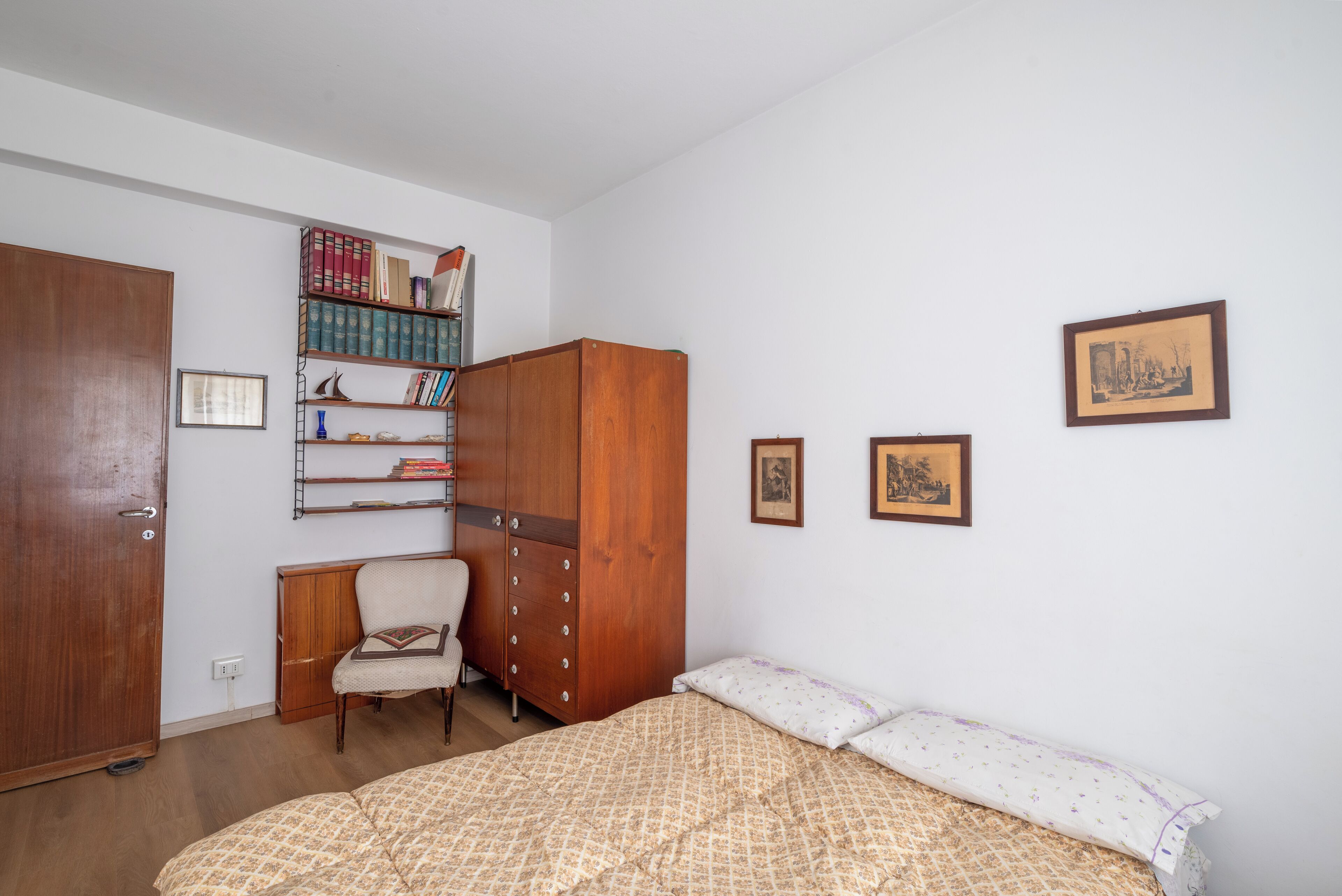 Een slaapkamer met een bed, een houten garderobe, een stoel, een boekenkast en ingelijste foto's aan de muur.