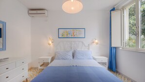 Une chambre à coucher comprenant un lit, deux tables de chevet, un climatiseur fixé au mur, un luminaire au plafond, une fenêtre avec des rideaux et un tableau encadré accroché au mur.