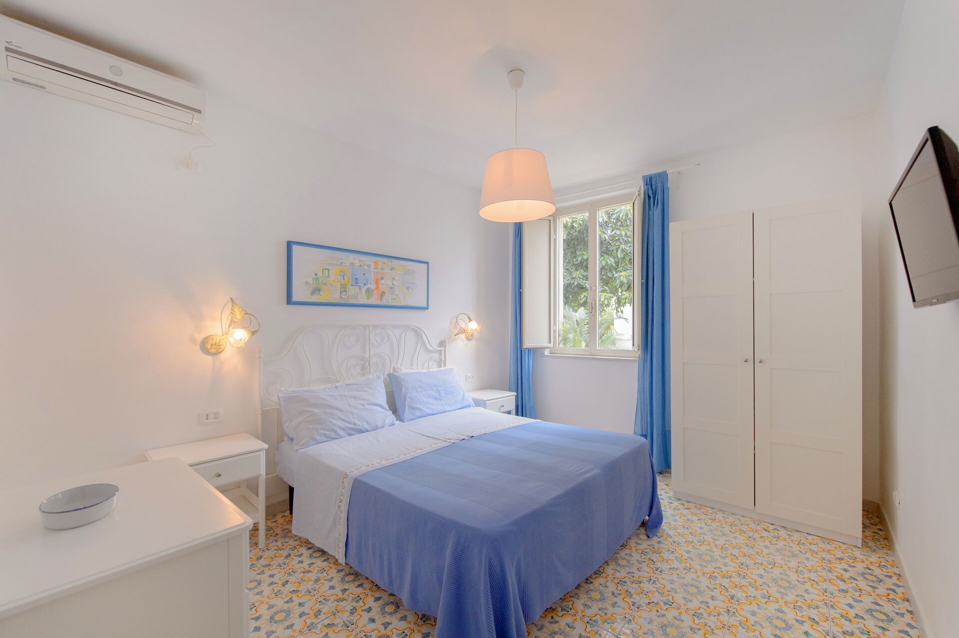 A bedroom with a bed, bedside tables, a window with curtains, a wall-mounted air conditioner, and a painting.