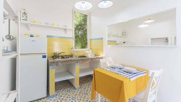 A compact kitchen with a gas stove, sink, and a window. There is a small dining table with a yellow tablecloth and a chair. The floor has patterned tiles.