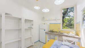 Une cuisine compacte avec des meubles blancs, un petit réfrigérateur, une cuisinière à gaz et une fenêtre donnant sur un espace verdoyant.