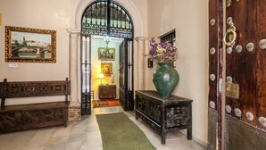 A hallway with a bench, a green vase with flowers, a wooden door with decorative metalwork, and a framed picture on the wall.