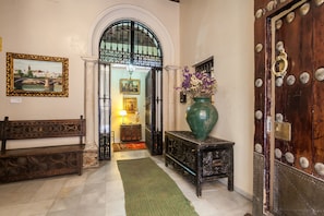 A hallway with a bench, a green vase with flowers, a wooden door with decorative metalwork, and a framed picture on the wall.