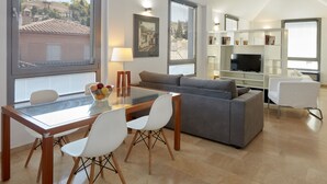 Un salon moderne avec une table à manger en verre, un canapé gris et des chaises blanches. On y trouve un téléviseur posé sur un meuble blanc, ainsi qu’un tableau accroché au mur.