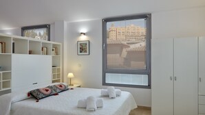 Une chambre moderne avec un grand lit, une armoire, une table de chevet avec une lampe, et une vue sur les bâtiments par la fenêtre.