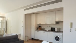 Une cuisine moderne avec des meubles blancs, un four à micro-ondes et un lave-linge. On y trouve une table à manger en verre, des chaises et un saladier rempli de fruits.