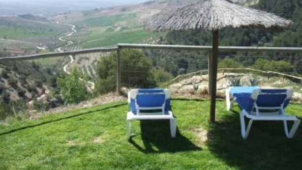Una terrazza con due poltrone e un ombrellone di paglia, con vista su una valle e una strada tortuosa, immersa nel verde.