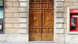 Une porte en bois à deux battants, surmontée d’un arc en pierre, avec une plaque de numéro et un distributeur automatique de billets rouge placé à droite.