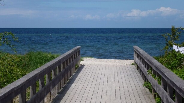 Une promenade en bois qui mène à une plage où l’eau est d’un bleu limpide et la mer est calme.