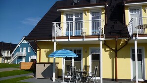 Un immeuble résidentiel de deux étages doté d’un balcon, d’un petit espace extérieur aménagé avec une table et des chaises, et d’un parasol à rayures bleues.
