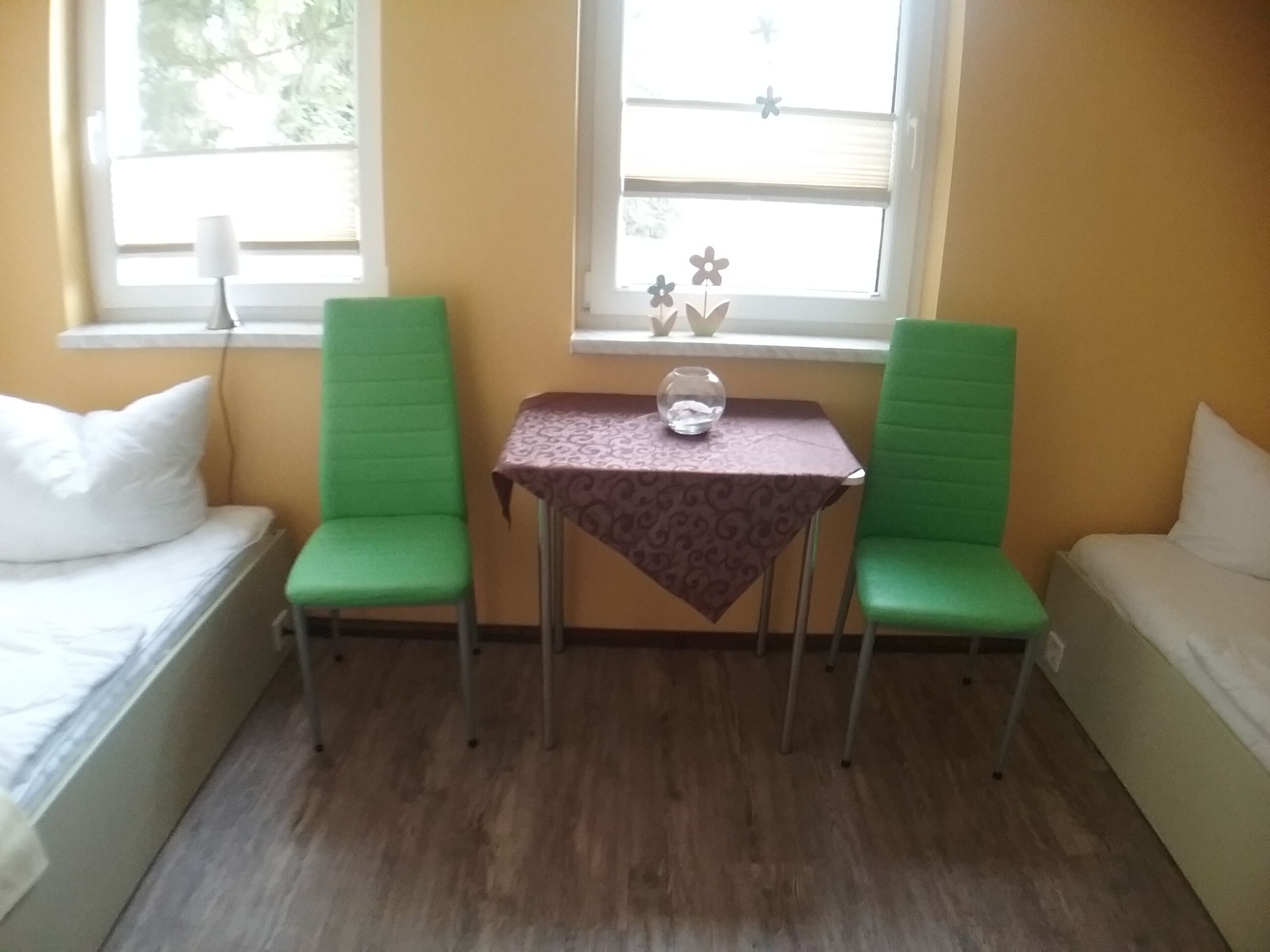 Ein kleiner Raum mit zwei grünen Stühlen, einem Tisch mit gemustertem Tischläufer und einem Fenster mit Dekoration.