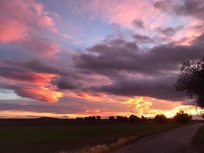 A rural landscape with a road curving through fields and trees, under a colorful sunset sky.