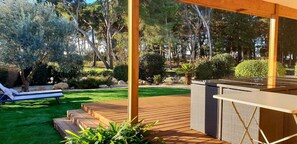 Une terrasse en bois dotée d’un bar intégré, d’un fauteuil de relaxation et d’une pelouse bien entretenue agrémentée d’arbres et d’arbustes.