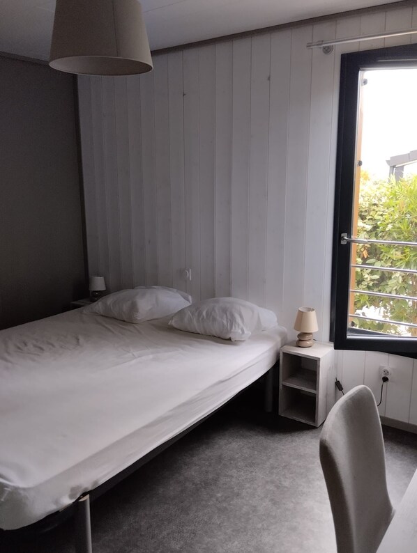 Un lit simple avec du linge de lit blanc, une table de chevet avec une lampe et une fenêtre donnant sur un paysage verdoyant.
