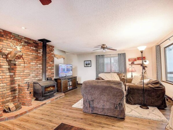 A living room with a brick fireplace, a television, a ceiling fan, and a large window. - Deck w Hammocks AIR Conditioning & EV Charger! Escape to the Woods - Flagstaff Rental Cabin (Flagstaff)