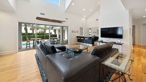 A spacious living area with a large sofa, a glass coffee table, and a kitchen area with a bar and a TV. - Time Out - Destiny East Lux Villa. Pool, Hot Tub, 2 Golf Carts 🛺, 6BR (Destin)