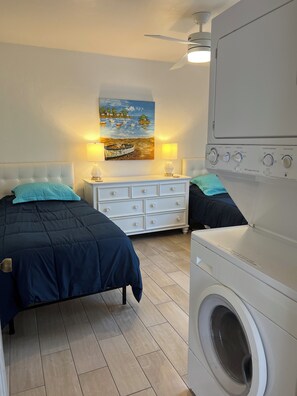 Un dormitorio con cama, cómoda, mesita de noche, lámpara, cuadro y lavadora.