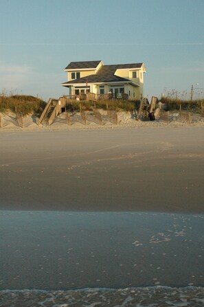 Una casa frente a la playa con una escalera que lleva a la entrada, rodeada de dunas de arena y vegetación costera.