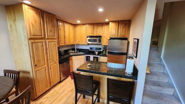 Una cocina con armarios de madera, microondas, estufa y refrigerador. Hay dos taburetes de bar y una silla de madera.