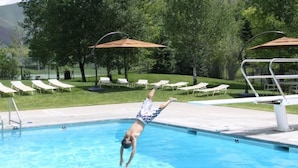 Una piscina con tabla de clavados, sillas de descanso y sombrillas.