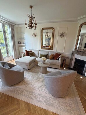 A modern living room with a sofa set, ottoman, and a round coffee table. There is a fireplace, a chandelier, and a large mirror on the wall.