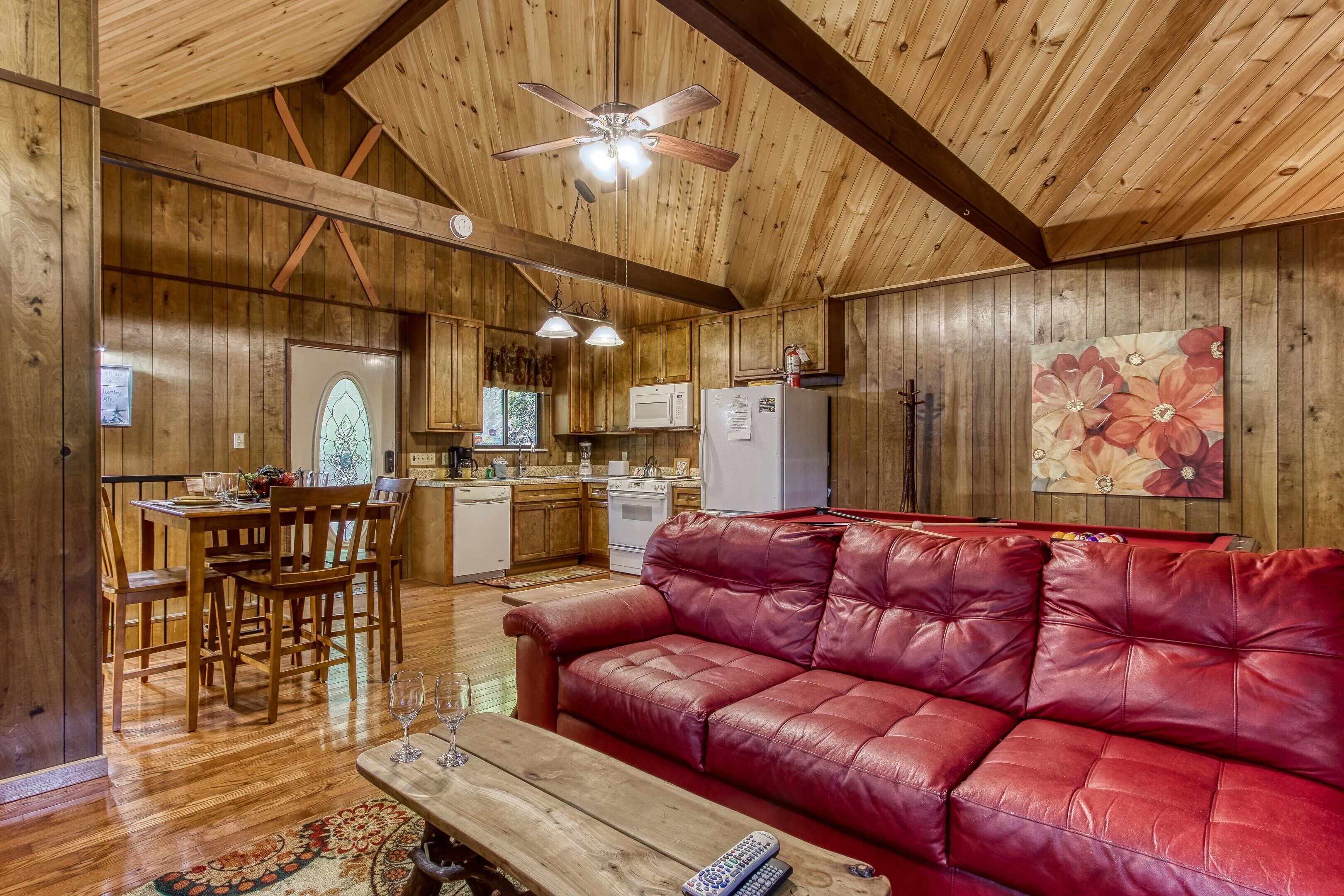 A wooden interior with a red leather sofa, wooden dining table, and a ceiling fan.