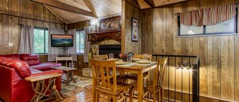 A wooden cabin interior with a dining table set for four, a red leather sofa, and a fireplace.