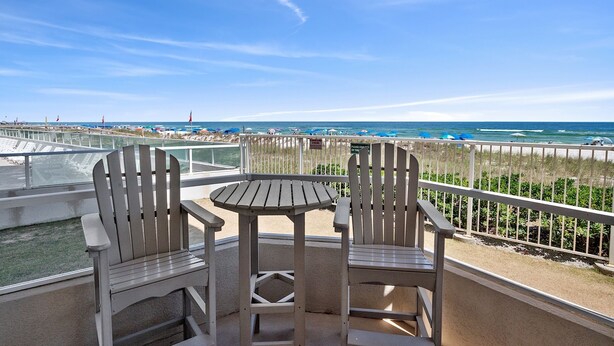 Un balcon avec deux fauteuils Adirondack et une petite table, donnant sur une plage parsemée de parasols et bordée d’une clôture.