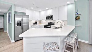 Une cuisine moderne avec des meubles blancs, des appareils en acier inoxydable, un grand îlot central doté de tabourets et un mur d’accent de couleur turquoise clair.