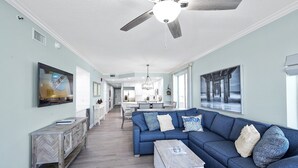 Un salon spacieux avec un canapé bleu, une table basse en bois et un ventilateur de plafond. On aperçoit une cuisine et un coin repas au fond.