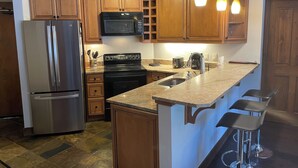 A kitchen with wooden cabinets, stainless steel appliances, a granite countertop, and bar stools. - Ski-In, Ski-Out, Corner Unit. Sleeps 4. Hot Tubs, Pool, 65" OLED TV. Near Base (Winter Park)