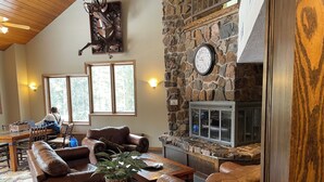 A cozy living room with leather sofas, a stone fireplace, a clock, and a person working at a table by large windows. - Ski-In, Ski-Out, Corner Unit. Sleeps 4. Hot Tubs, Pool, 65" OLED TV. Near Base (Winter Park)