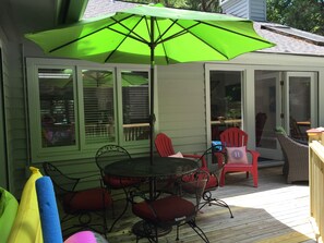 A covered patio area with a table and chairs, a green umbrella, and a view of the house through the windows. - Celebrate at the Beach Thanksgiving! Private Pool and Free Bikes! (Hilton Head Island)