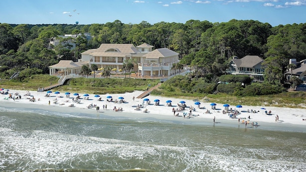 A beachfront property with a large house, a sandy beach with umbrellas, and a forested hill in the background.