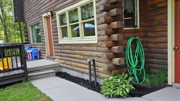 A log cabin with a front porch, a small garden area, and a hose coiled against the wall.